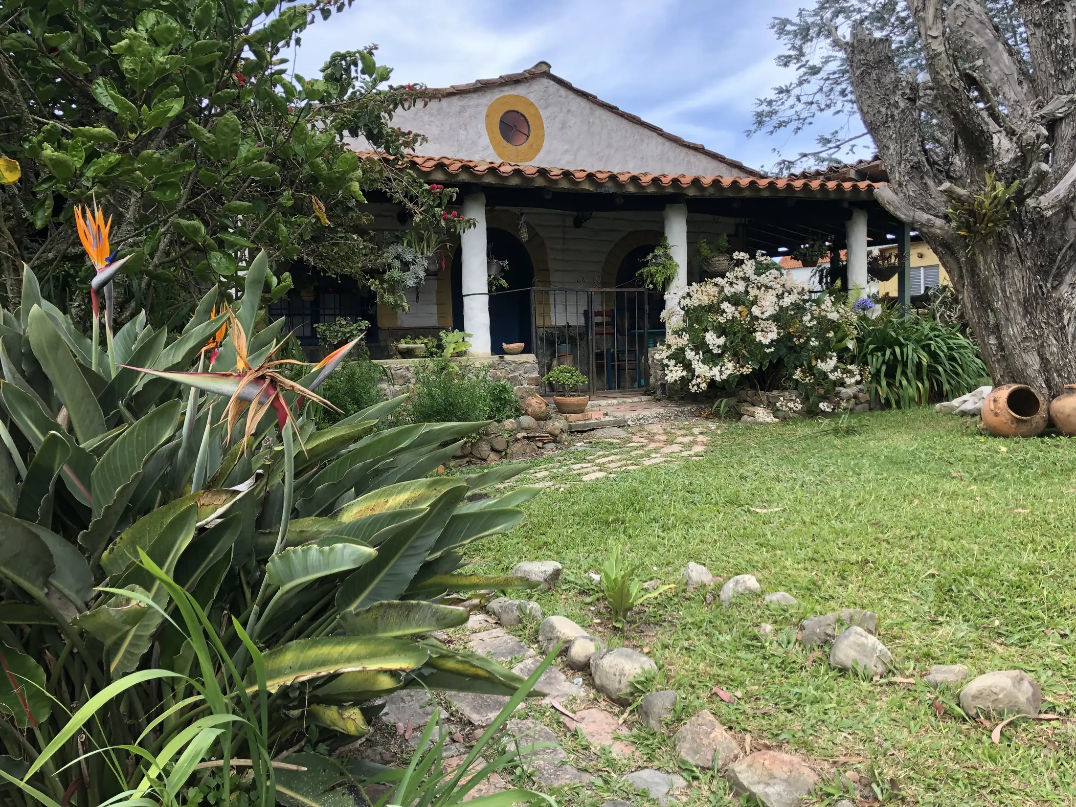 La casita: fachada blanca con techo de teja, aro amarillo en el frontón y jardín con flores